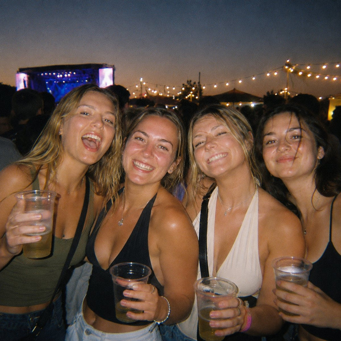 Four friends with drinks in a festival crowd at dusk, warm grainy tones
