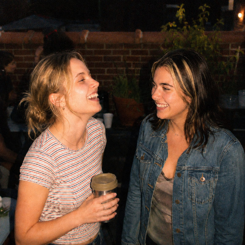 Two friends laughing outdoors at golden hour, one holding a drink