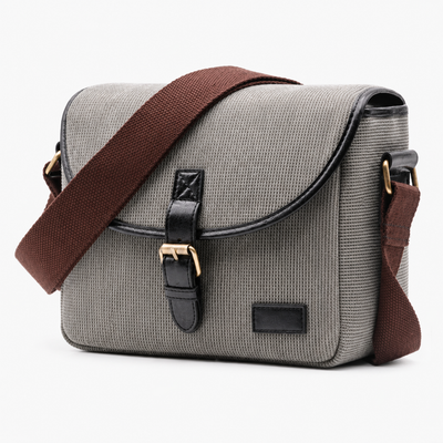 A gray canvas messenger bag with a brown fabric shoulder strap, black leather trim, and gold-tone hardware, shown on a plain white background.