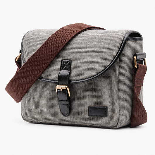 A gray canvas messenger bag with a brown fabric shoulder strap, black leather trim, and gold-tone hardware, shown on a plain white background.