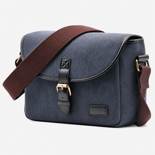 A navy blue canvas messenger bag with a brown fabric shoulder strap, black leather trim, and gold-tone hardware, shown on a light gray background.