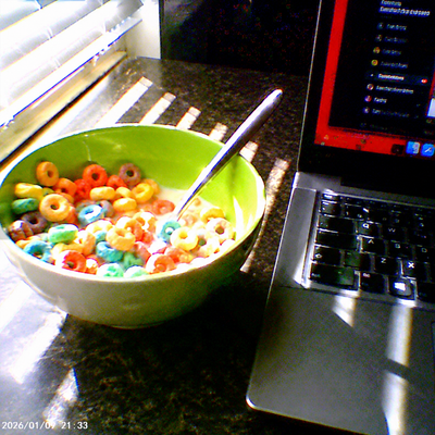A green bowl filled with colorful cereal and a spoon sits on a dark countertop next to an open laptop computer.