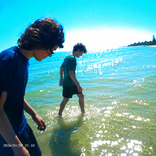 Two people wading in shallow ocean water under a bright blue sky, with sunlight reflecting off the water surface and a small shoreline visible in the distance.