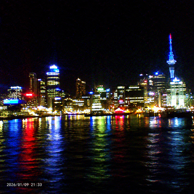 Nighttime city skyline with illuminated skyscrapers and colorful reflections on water, photographed from across the water with a timestamp in the lower-left corner.