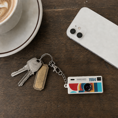 A retro-style camera keychain with keys and a blank tag, styled on a dark wooden table next to a coffee cup and a smartphone.